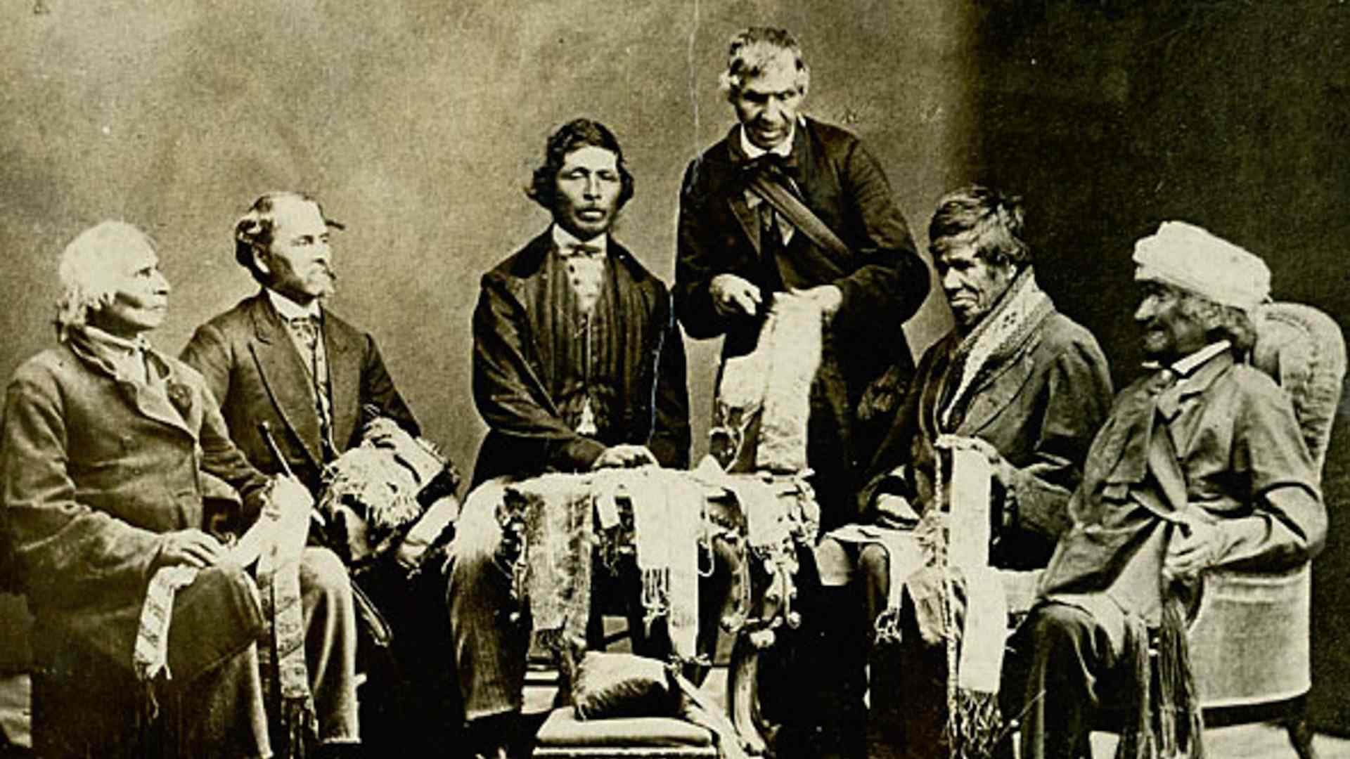 Five Native American chiefs, dressed in western clothes, sitting in a semi-circle holding belts made of wampum beads