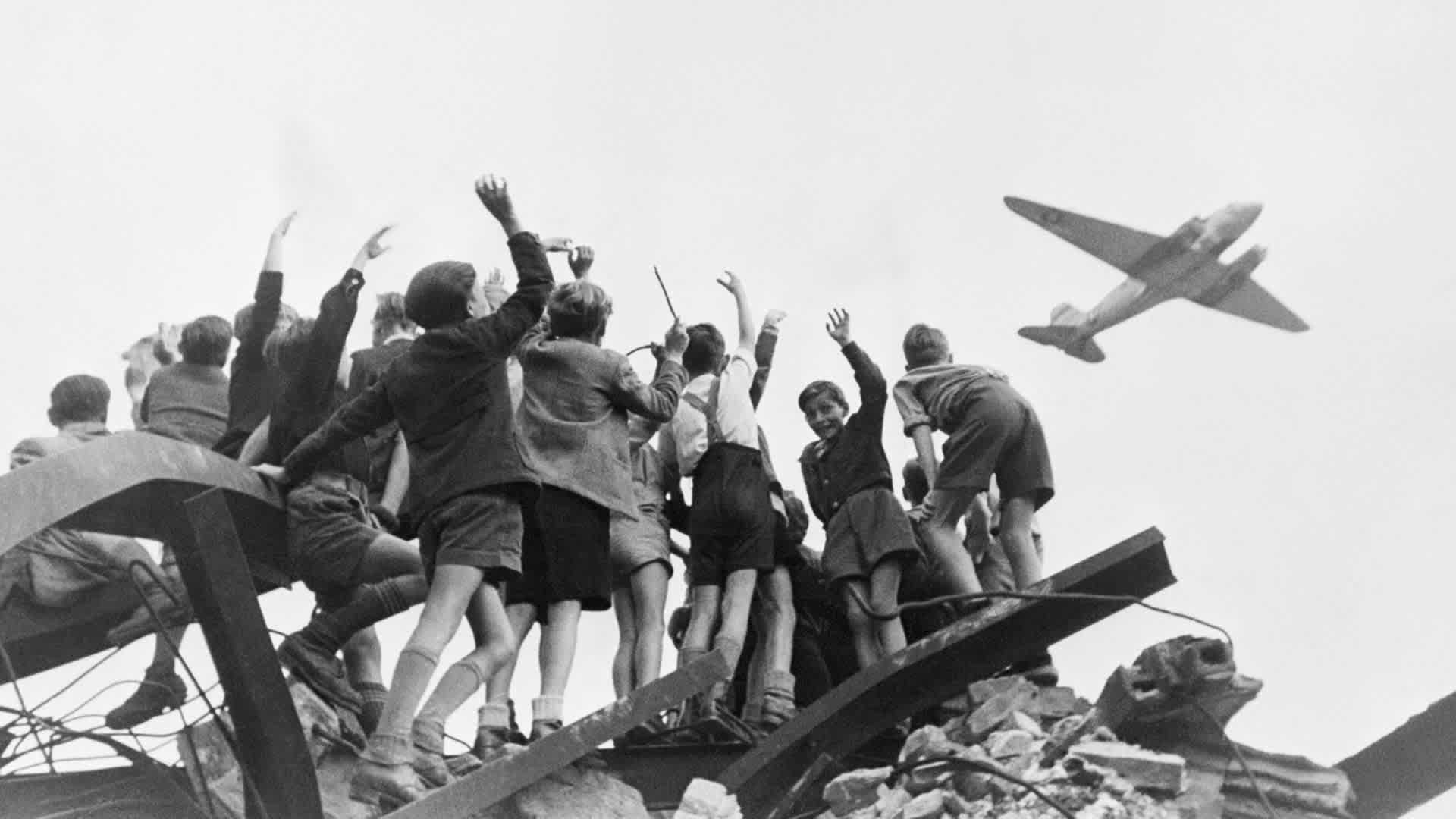 Berlin Airlift - A group of German children stand atop building rubble, cheering a U.S. cargo airplane as it flies over a western section of Berlin in January 1948.