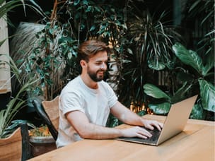 A cheerful man, sitting at a desk, working on his laptop, with plants in the background