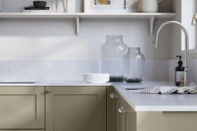 Wardley French clay kitchen cabinetry with steel knobs, steel tap, white marbled worktop and matching backsplash. An open shelf on the wall above cabinets with ceramics and a vase of flowers.
