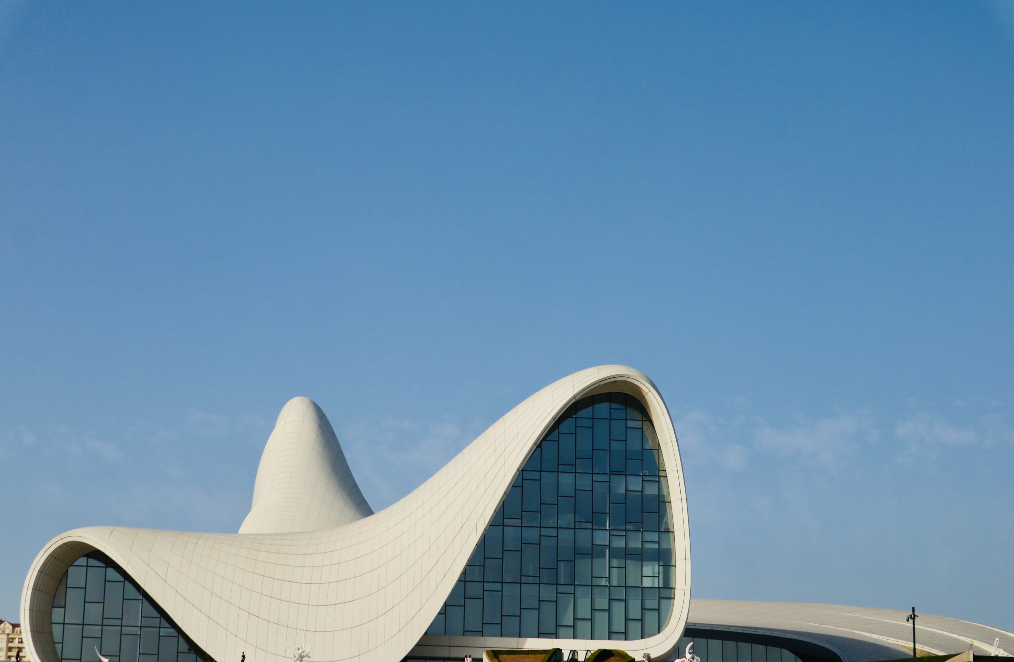 A futuristic, curvilinear building with a white, flowing roof and large, blue-tinted glass windows stands against a clear, blue sky in Azerbaijan. The design features smooth, wave-like contours, creating a modern and dynamic architectural form.