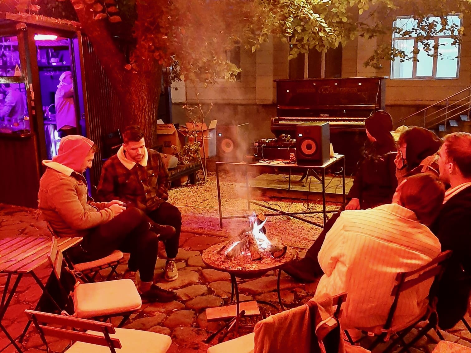 A group of people sitting around a campfire at night in a quaint courtyard, the warm lighting creating a cozy atmosphere reminiscent of Sighnaghi, the City of Love. As you soak in the ambiance, imagine the gentle melodies playing from a piano and audio equipment set up in the background. This charming town offers not just breathtaking views and historic architecture but also enchanting experiences like evenings filled with music and camaraderie. Whether you're exploring its cobblestone streets or enjoying moments like this under the Georgian sky, Sighnaghi promises unforgettable memories.