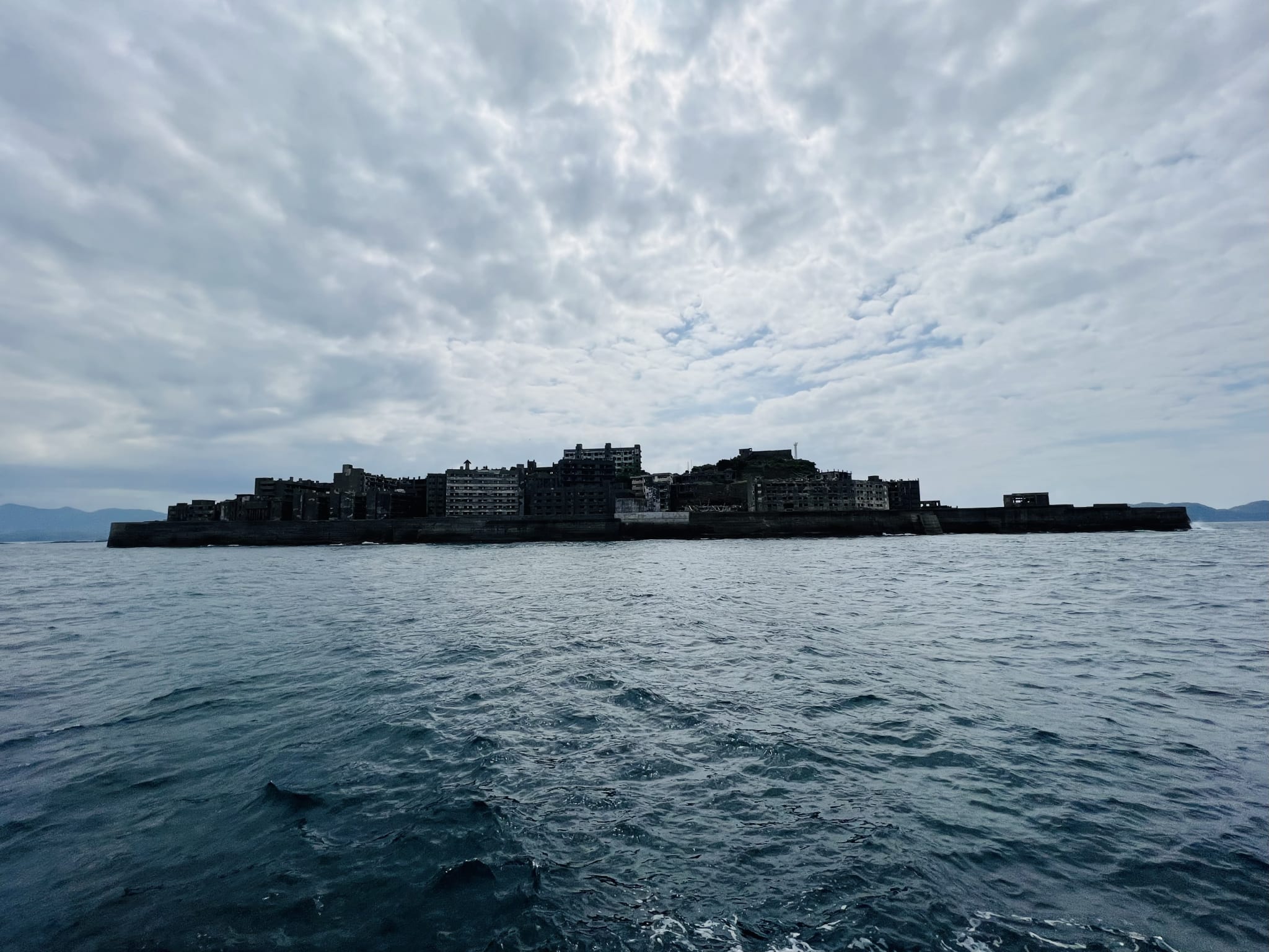 Silhouette of an abandoned island with dilapidated buildings under a cloudy sky, surrounded by dark, choppy waters. This mysterious landmark could be a hidden gem for those looking to explore Kyushu beyond the typical travel guide.