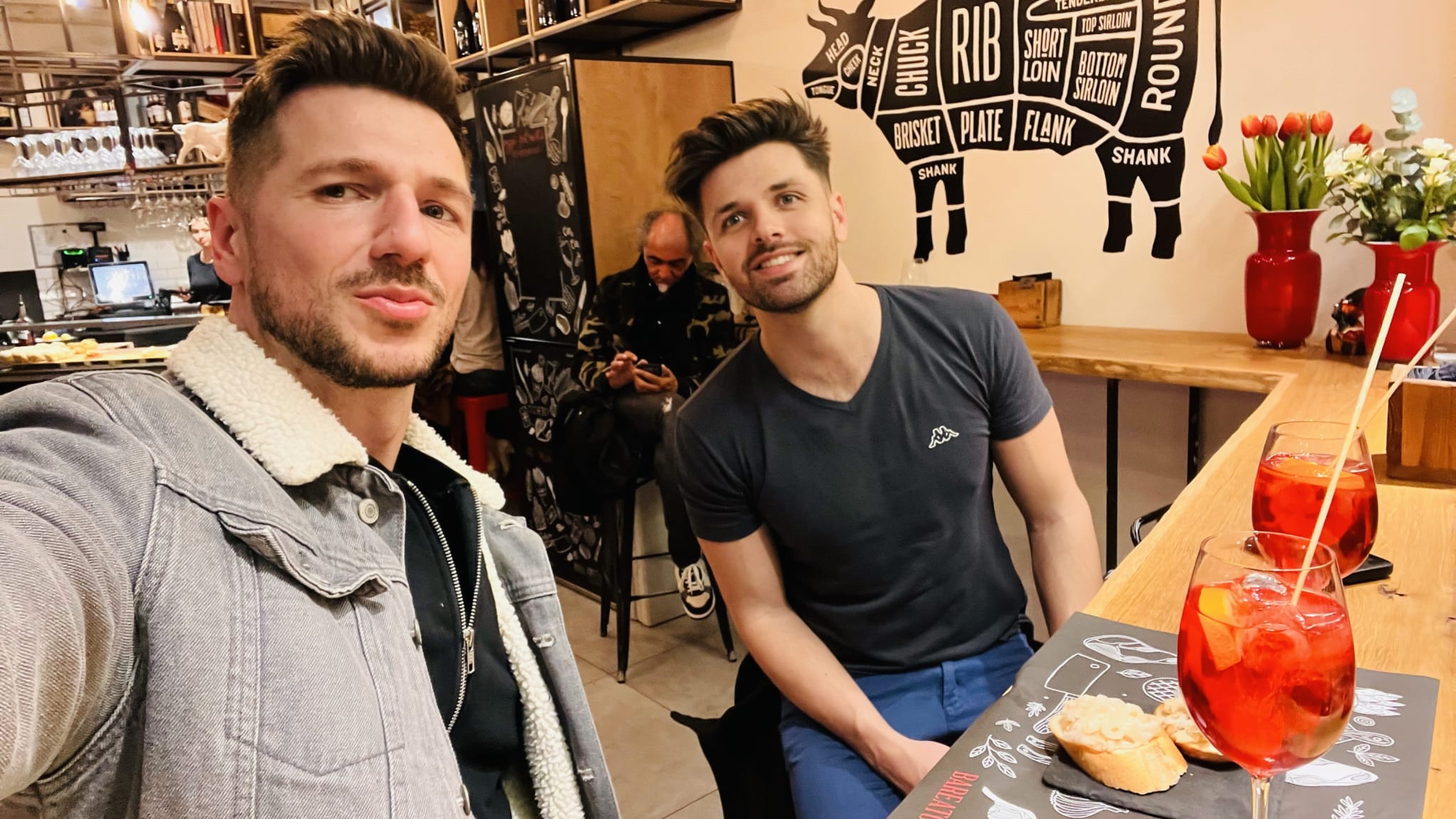 Two people enjoying a meal in Venice sit at a restaurant table adorned with drinks and food. One person in a denim jacket holds up a phone for a selfie, celebrating their moment in this captivating city known for its gay-friendly ambiance. The other looks on with joy as the charming decor of the eatery enriches their travel memories in gay Venice (or gay Venezia).