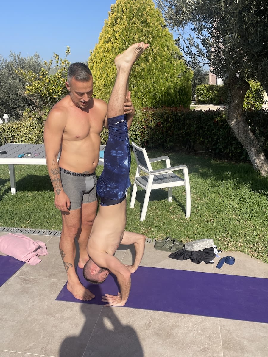 Man assisting another man in a headstand pose during a yoga session in a garden.