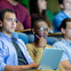 A diverse group of college students are sitting in a university classroom. They are taking notes while listening to their professor (located offscreen) during the lecture.