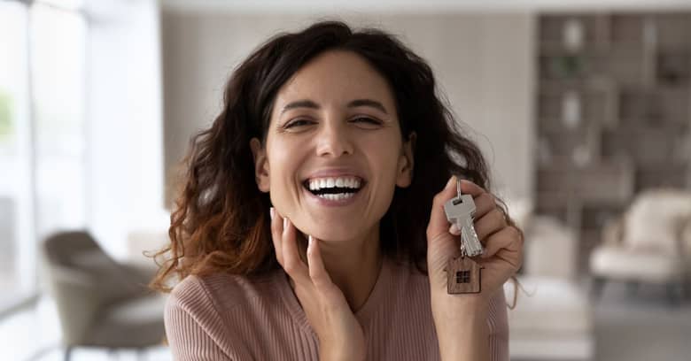 Smiling woman holding a set of house keys with a small wooden house keychain, expressing excitement.